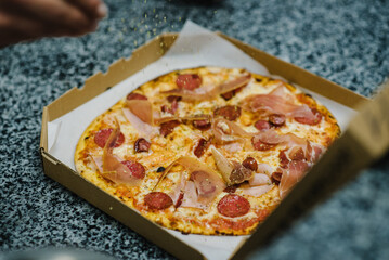 Chef is sprinkling fresh oregano over a made pizza. Baked pizza in a box. Traditional Italian pizza with meat, prosciutto, salami, cheese. Food delivery. Self-pickup or home delivery. Top view.