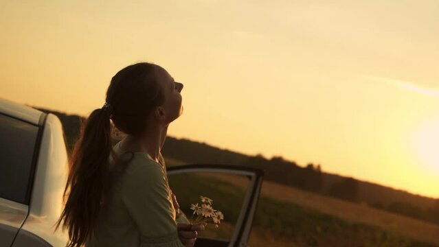 Woman stops on the side of the road to admire the sunset, leaning on the car door. 