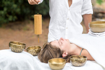 A man wearing in white holding a bamboo Koshi chime musical instrument during sound healing therapy with tibetan singing bowls over young woman during sound massage outdoor, selective focus