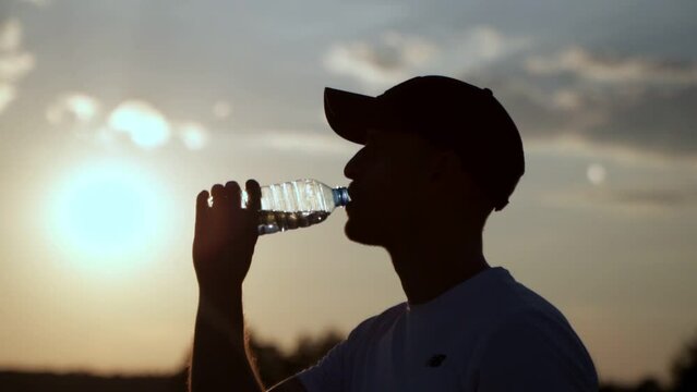 Silhouette of a fitness man in a cap drinking water from a bottle. 