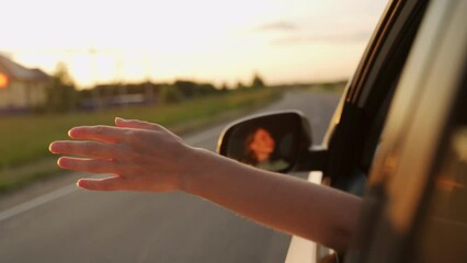 A woman with long hair is sitting in the driver's seat of a car, stretching her arm out of the window and catching the glare of the setting sun. A free woman traveling by car catches the wind with her