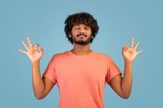 Peaceful indian guy meditating over blue stdio background
