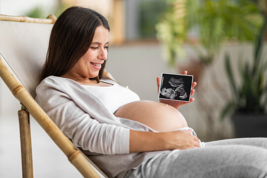 Pregnant Woman Showing Baby Ultrasound Photo While Relaxing In Chair At Home