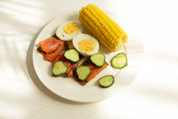Breakfast plate on white texture background. Fish, eggs, cucumbers and corn.