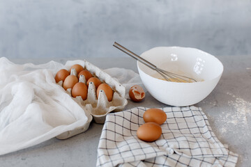 A tray with eggs and a white deep plate with a whisk on the kitchen table on a grey background.