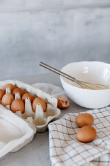 A tray with eggs and a white deep plate with a whisk on the kitchen table on a grey background.