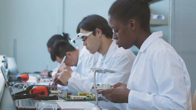 Young African American female computer engineer and her colleagues examining circuit board at desk in laboratory, entering data information on computers