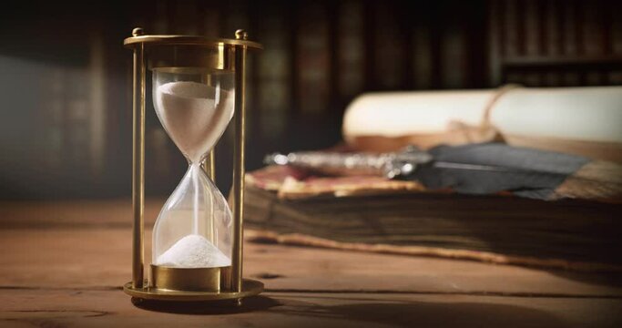 sand running through the hourglass on old wooden table with vintage books and quill pen. time is ticking
