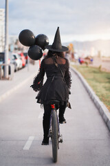 woman in witch costume with black balloons riding a bicycle on the street, back view