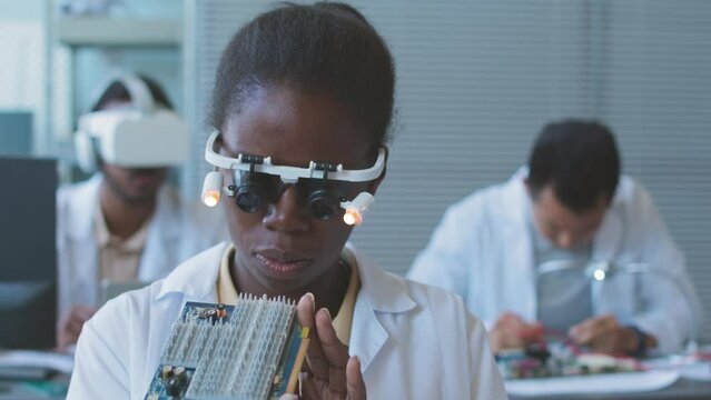 Concentrated young Black woman in headband magnifier examining motherboard while working with colleagues in computer engineering lab