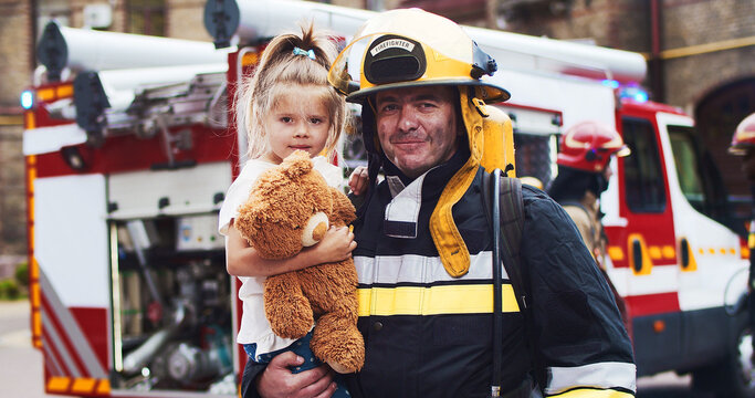 Portrait of caucasian handsome fireman in helmet and gull equipment holds girl with teddy bear in arms and looking into camera smiling. The concept of saving lives, heroic profession, fire safety