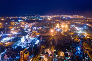 Obraz premium Aerial view of steel plant at night with smokestacks and fire blazing out of the pipe. Industrial panoramic landmark with blast furnance of metallurgical production. Concept of environmental pollution