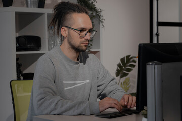 Fototapeta premium A European man with dreadlocks and glasses works at a computer in an office