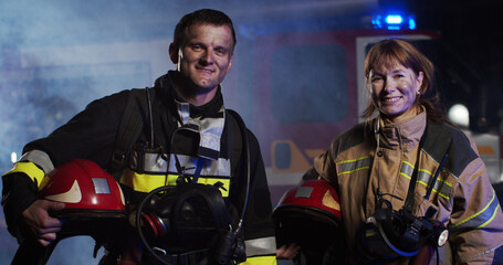 Portrait caucasian female and male fireman in gull equipment holding helmet in hands looking at camera and smiling near fire truck in smoke at night. Concept of saving lives, fire safety