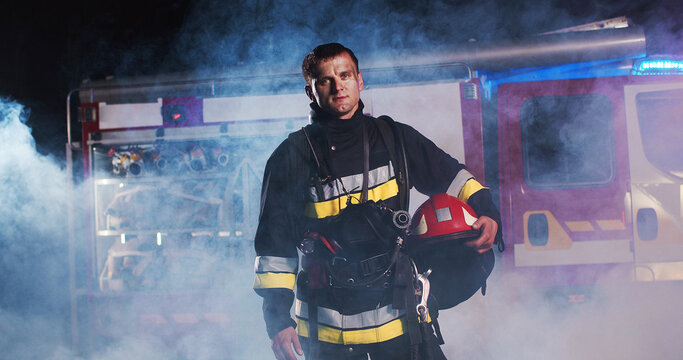 Middle plan of tired Caucasian young man fire fighter taking off helmet and walking out from fire at the red truck and looking into camera. The concept of saving lives, heroic profession, fire safety