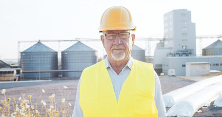 Portrait of industrial senior engineer in glasses with security hat and lifevest standing next to...