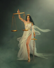 Portrait fantasy woman Greek goddess of justice Themis holding scales and sword in hands. White silk vintage dress old antique style flies waving in wind. Blind girl queen eyes blindfolded. art photo