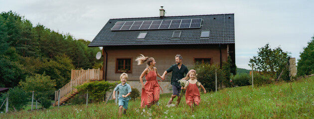 Happy family running near their house with solar panels. Alternative energy, saving resources and sustainable lifestyle concept. - obrazy, fototapety, plakaty