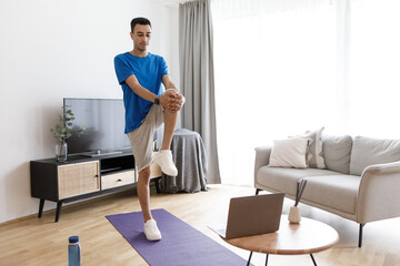 Home workout routine. Young arab man stretching and doing standing knee-to-chest exercise, using laptop computer
