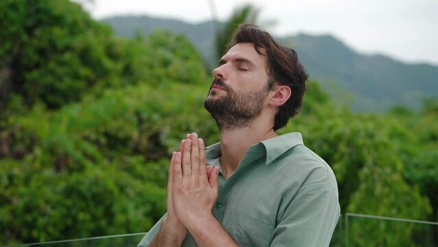 Peaceful bearded young man raises his head to the sky, saying prayers while praying to God, secluded in mountains, feeling harmony and connection with nature. Loneliness, mindfulness, spiritual growth