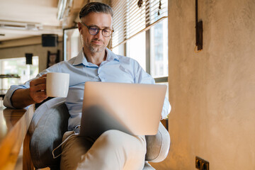European grey man drinking coffee while working with laptop at office