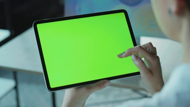 Back shot of unrecognizable young Caucasian woman holding electronic tablet, touching green screen, browsing. View of girl using gadget on blurred background. Indoors