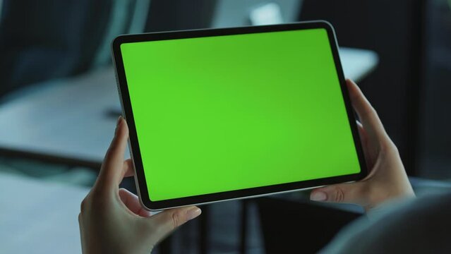 View of hands of young Caucasian woman using tablet with green screen. Shot of girl holding gadget in office. Blurred background of workplace. Indoors