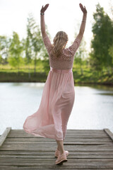 A beautiful woman in a flowing pink dress stands on a wooden bridge with her back to the camera.