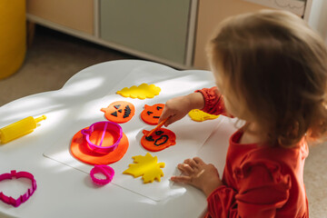 A little girl playing with playdough funny and angry pumpkins and autumn leaves for the holiday of Halloween. Sensory play for toddlers. Holiday Art Activity for Kids.