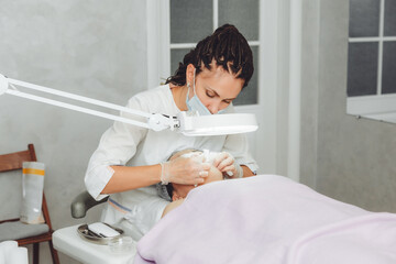 A cosmetologist cleanses the face under a cosmetic magnifying glass. The concept of cosmetology. A cosmetologist performs a facial cleansing procedure with problem skin.