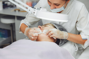 A cosmetologist cleanses the face under a cosmetic magnifying glass. The concept of cosmetology. A cosmetologist performs a facial cleansing procedure with problem skin.