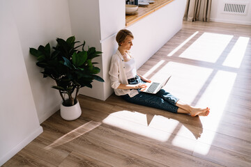 Concentrated female freelancer working on laptop while sitting on floor in apartment