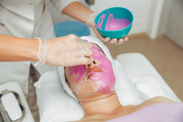 Obraz na płótnie Canvas The process of applying an alginate face mask. cosmetologist holding a spatula. A woman lying on a couch in the cosmetologist's office. Facial skin care in a beauty salon