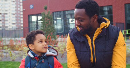 Close up of cheerful dad and son sitting outdoors near school and chatting in good mood. Joyful...