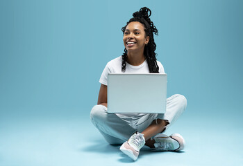 Happy young woman sitting on the floor with crossed legs and using laptop on blue background. - 533394465