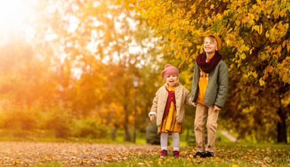 Fototapeta premium Autumn park children boy and girl stay on path, laugh and looking at camera in fall golden leaves meadow October season. Kids wearing knitted casual stylish clothes. Positive emotion. Copy space