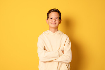 Happy young caucasian boy in casual outfit with arms crossed isolated over yellow background
