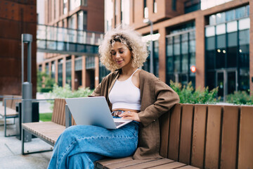 Smiling ethnic woman sitting on bench and looking at laptop screen in street