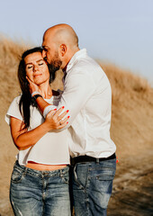 Happy smiling  Couple walking at Sand Dunes near the Beach.  Young happy Bearded muscular  man  in White shirt kissing and hugging beautiful woman at sunser on a beach