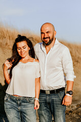 Happy smiling  Couple walking at Sand Dunes near the Beach.  Young happy Bearded muscular  man  in White shirt kissing and hugging beautiful woman at sunser on a beach