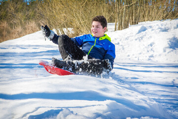 In winter, in a forest, a boy lying on his stomach slides down a hill on a plastic plate. He is...