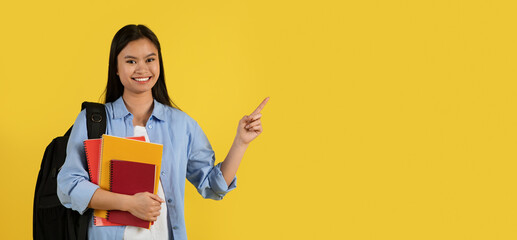Smiling young chinese female student in casual with backpack and many books pointing finger at empty space