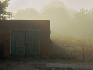The ruined facade of a garage with nothing behind but overgrown wasteland, the unreal, magical feel heightened by the thick mist and dawn sunbeams.
