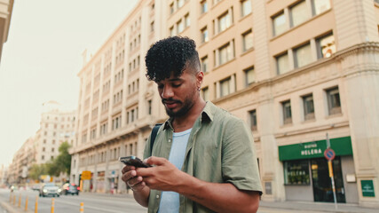 Young smiling man with beard dressed in an olive color shirt uses phone map app on the old city background