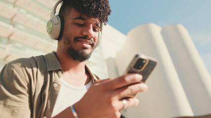 Fototapeta premium Close up, young male student dressed in an olive color shirt sits outside next to the university, listens to music on headphones, selects tracks on his phone