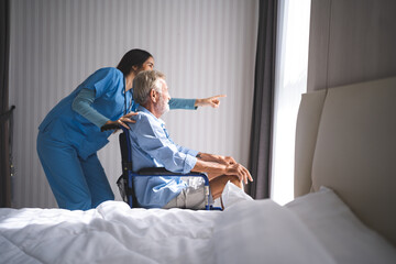 Full length portrait of Asian young female nurse assisting old senior man in wheelchair to looking at retirement home, carrying about disabled grey haired man in wheelchair, health insurance concept