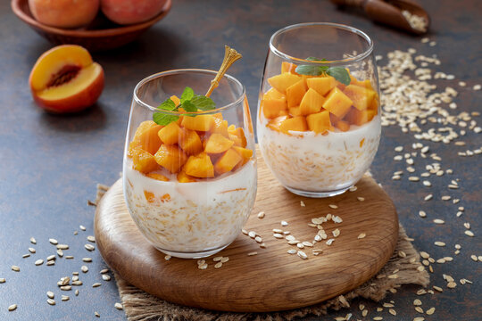 Peach Overnight Oats. Healthy breakfast or dessert made with oat grain soaked for the night in milk or yogurt topped with cubes of fresh peaches, served in a clear glasses. Dark, selective focus