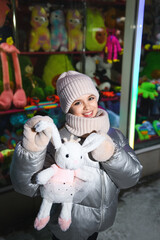 Girl holding a rabbit toy by the ears against the background of a showcase with trendy toys at night. Smiling girl in warm clothes: a jacket, a hat, a scarf. Holidays theme