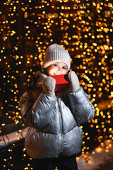 Girl and red gift box with Christmas lights. Smiling girl in a hat, scarf looking at the camera at night with blurry lights. Holidays theme