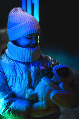 Girl with a dog toy against the background of a showcase with toys with neon illumination. Smiling girl in warm clothes: a jacket, a hat, a scarf at night. Holidays theme